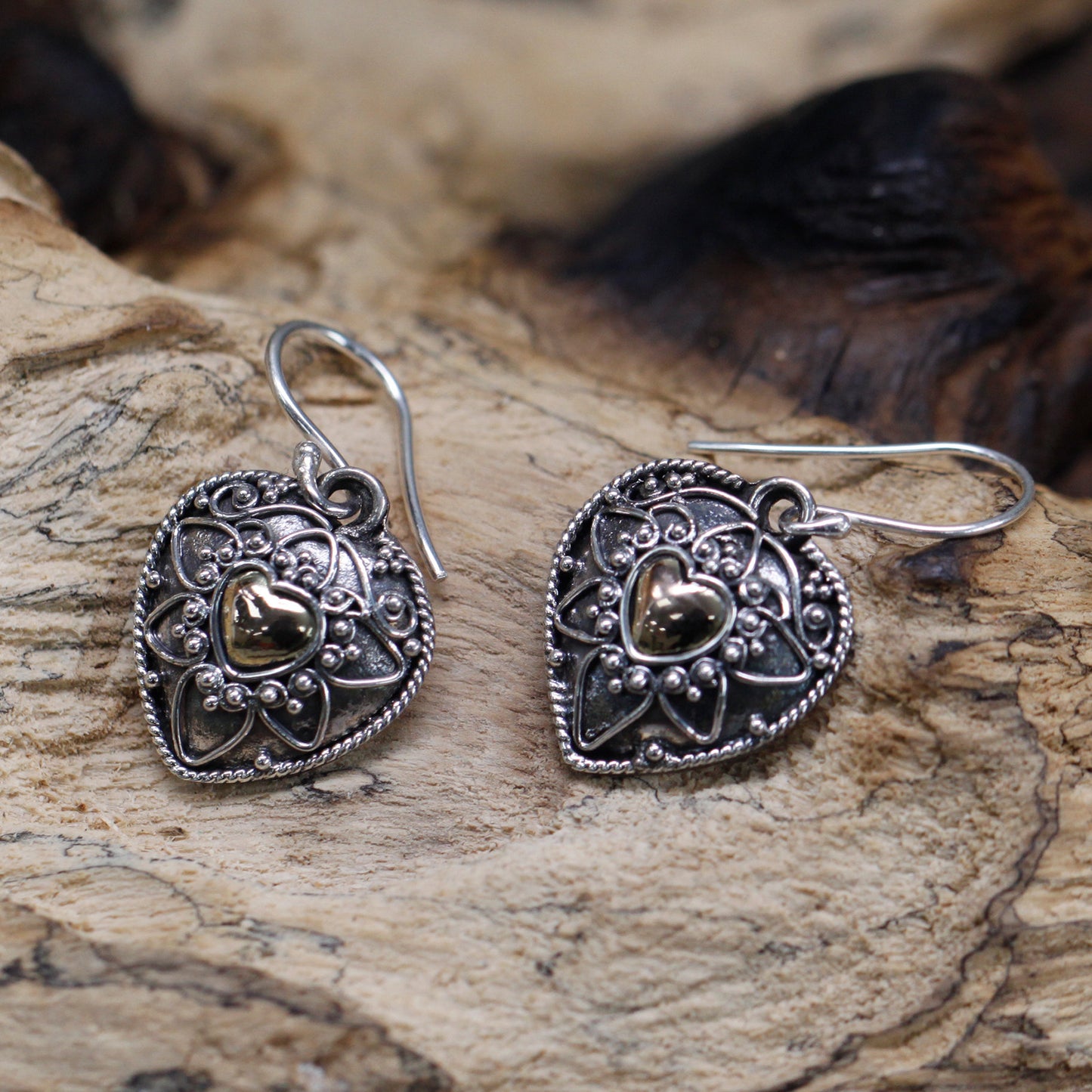 A pair of handmade silver and gold heart-shaped Mandala earrings resting on a textured wooden surface, highlighting the fine craftsmanship and ornate details