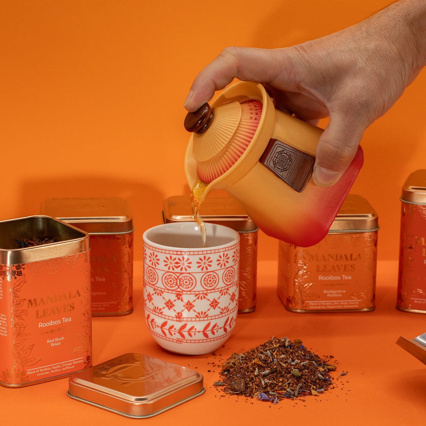 Red Bush Relax Blend Artisan Tea - Tinned - 50g, beside a patterned cup as red tea is poured from a yellow pot by Quintessential Holistic Health Ltd