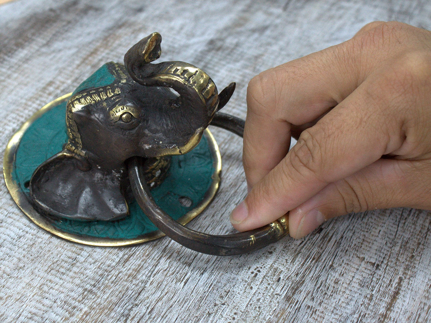 Brass Door Knocker - Elephant's Head, 8 × 9.5 × 13cm with curled trunk and a hand lifting the dark ring over a turquoise plate by Quintessential Holistic Health Ltd