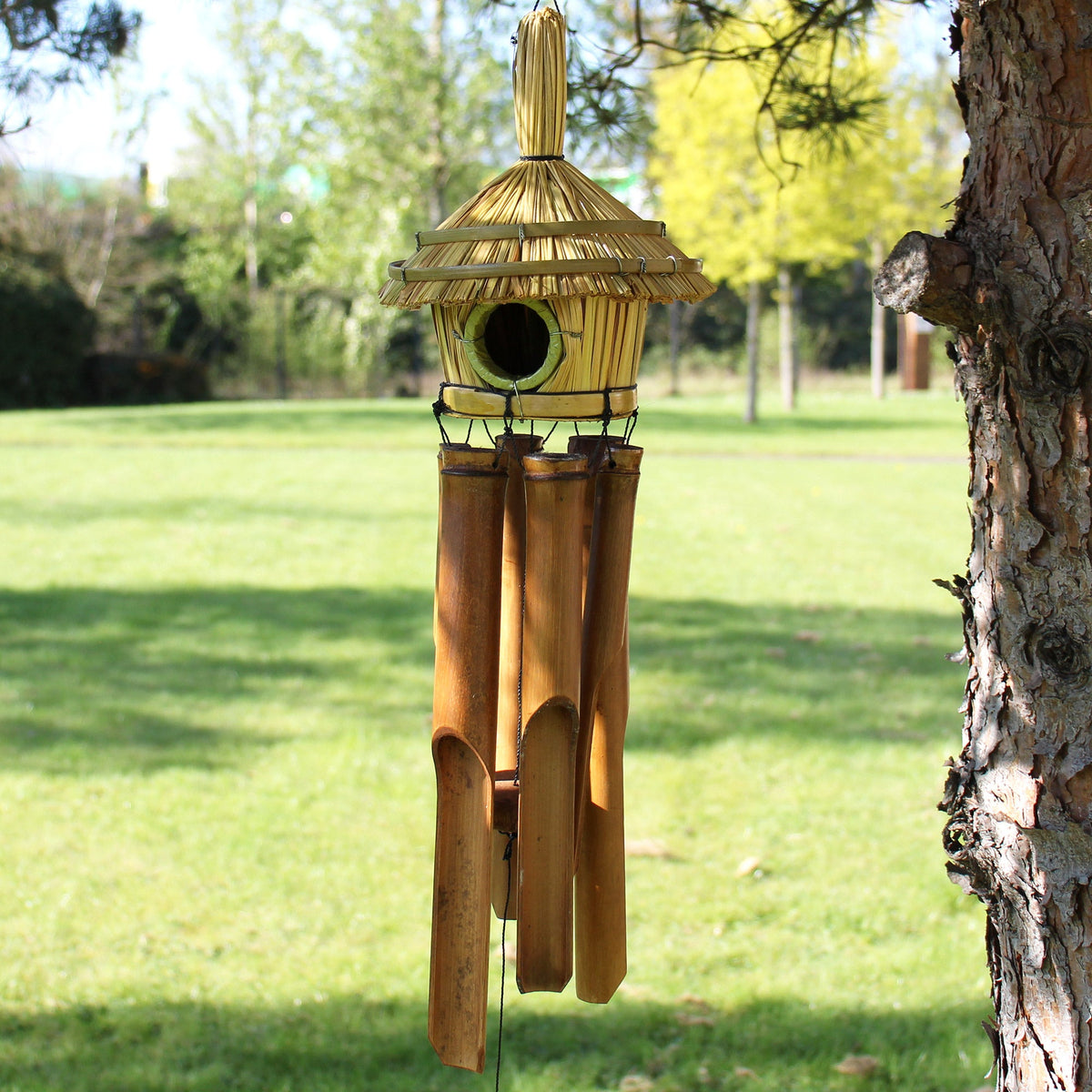 Handcrafted Seagrass & Bamboo Wind Chime Bird Box - Medium Round, hanging beside a tree trunk with a pointed roof and dark circular opening, by Quintessential Holistic Health Ltd