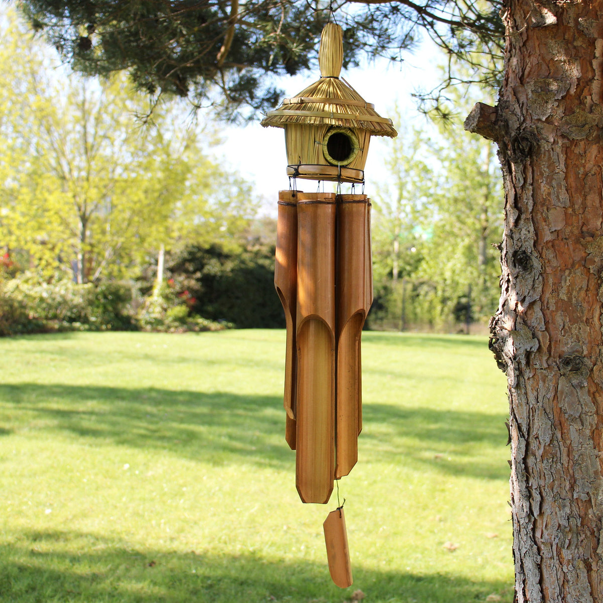 Handcrafted Seagrass and Bamboo Wind Chime Bird Box - Large - Round beside a tree trunk with woven roof and hanging tubes by Quintessential Holistic Health Ltd