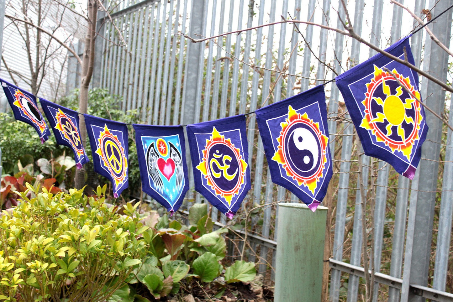 Handmade Batik Seven-Flag Rayon Banner - Love in the Centre hanging above plants with seven purple pointed flags and multicoloured emblems, by Quintessential Holistic Health Ltd