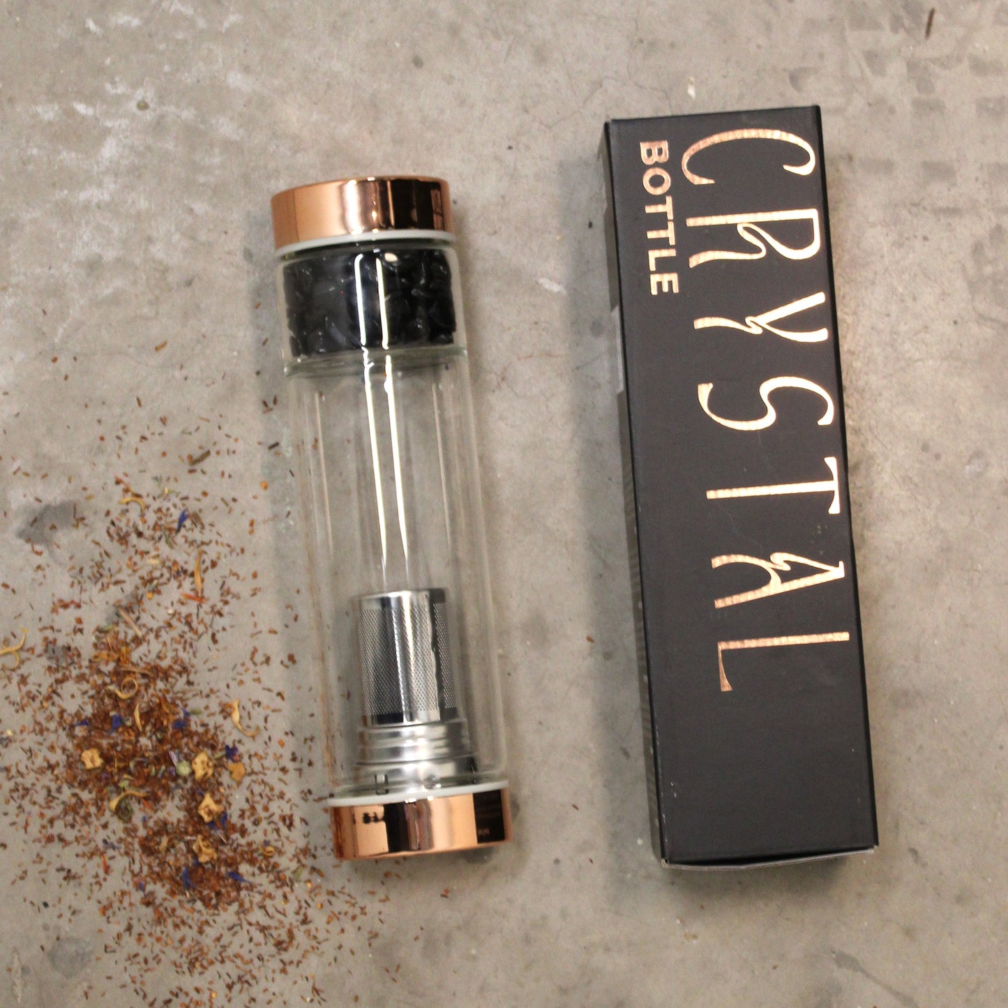 Rose Gold and Crystal Glass Tea Infuser Bottle with Rose - 500ml laid flat beside a black box and scattered loose leaves by Quintessential Holistic Health Ltd