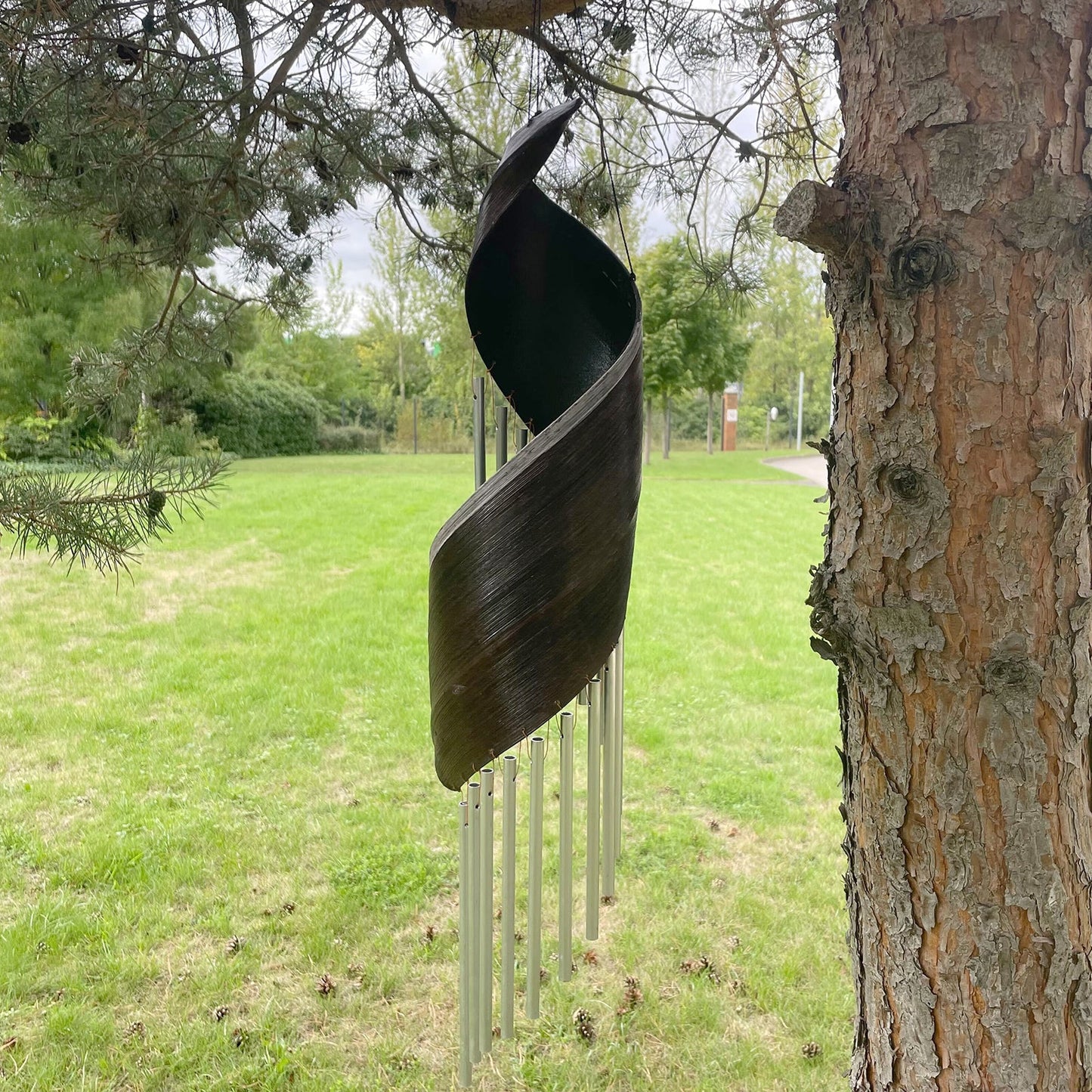 Handcrafted Coconut Leaf Wind Chime - Chocolate hanging beside a tree with a spiralled dark leaf and silver-tone tubes by Quintessential Holistic Health Ltd