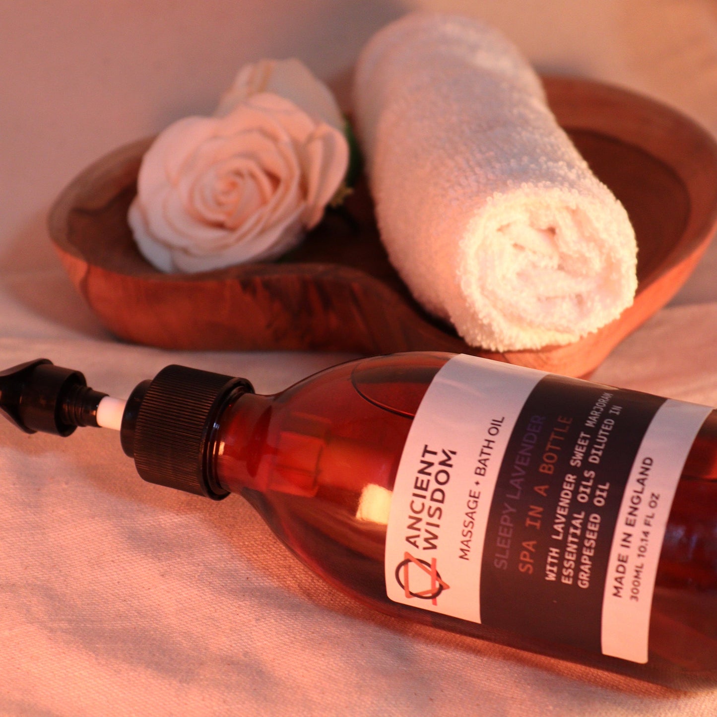 Sleepy Lavender 300ml Aromatherapy Massage & Bath Oil with Pump lying beside rolled towels and a pale rose by Quintessential Holistic Health Ltd