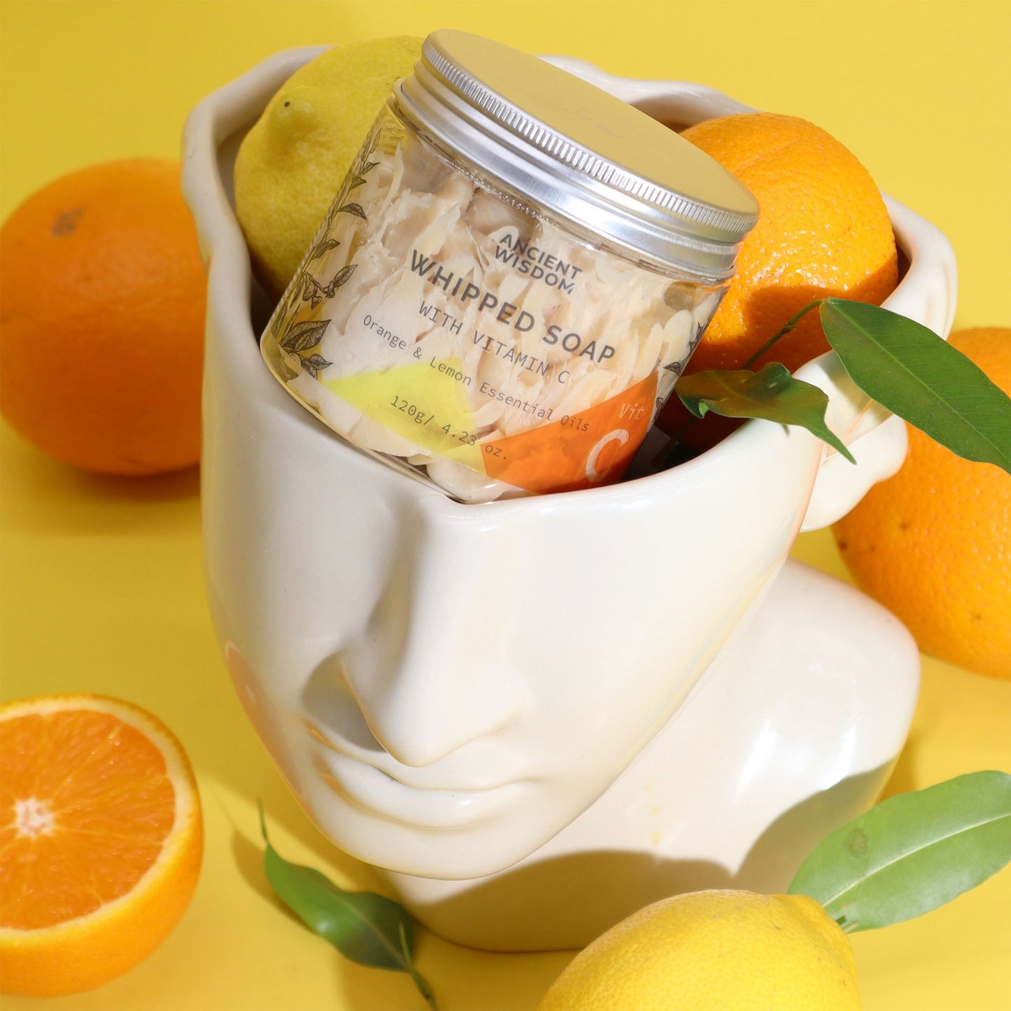 Vitamin C Aromatherapy Whipped Soap 120g, tilted jar in a white face-shaped pot surrounded by oranges by Quintessential Holistic Health Ltd