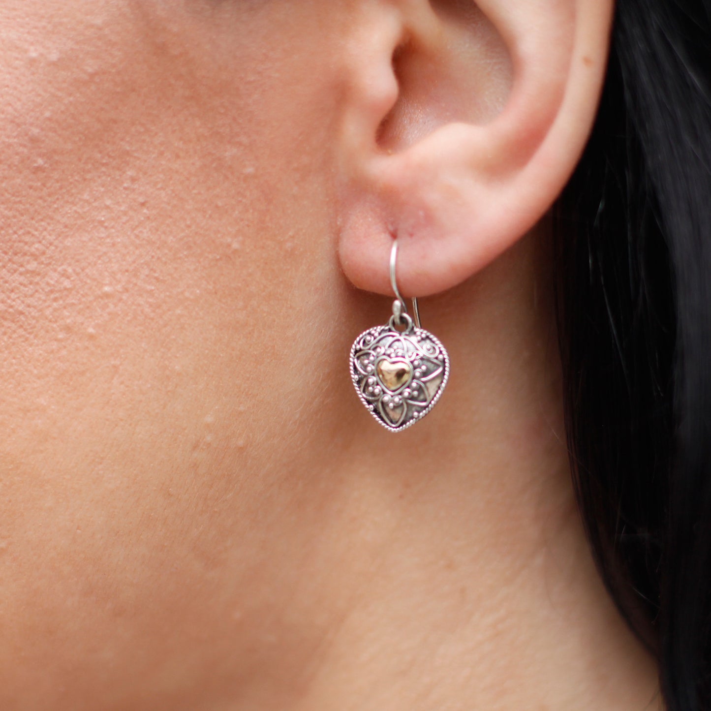A silver and gold handmade earring in the shape of a mandala heart, worn on an ear, showcasing its size and elegance