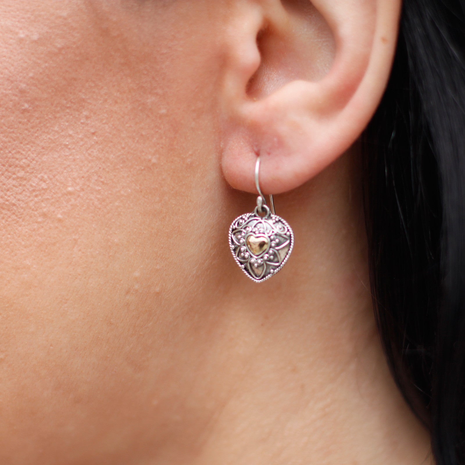 A silver and gold handmade earring in the shape of a mandala heart, worn on an ear, showcasing its size and elegance