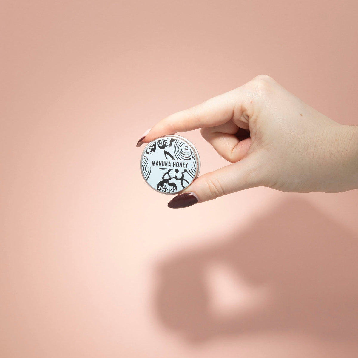 Hand-Poured Vegan-Friendly Natural Lip Balm - Manuka Honey, pale blue round tin held between two fingers against a peach backdrop, by Quintessential Holistic Health Ltd