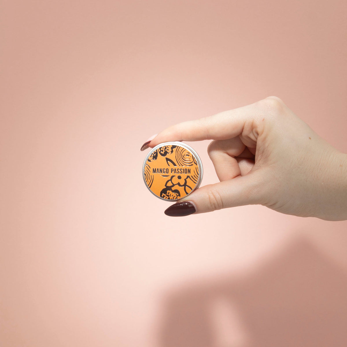 Hand-Poured Vegan-Friendly Natural Lip - Mango and Passion Fruit, orange round tin held between two fingers against a peach backdrop, by Quintessential Holistic Health Ltd