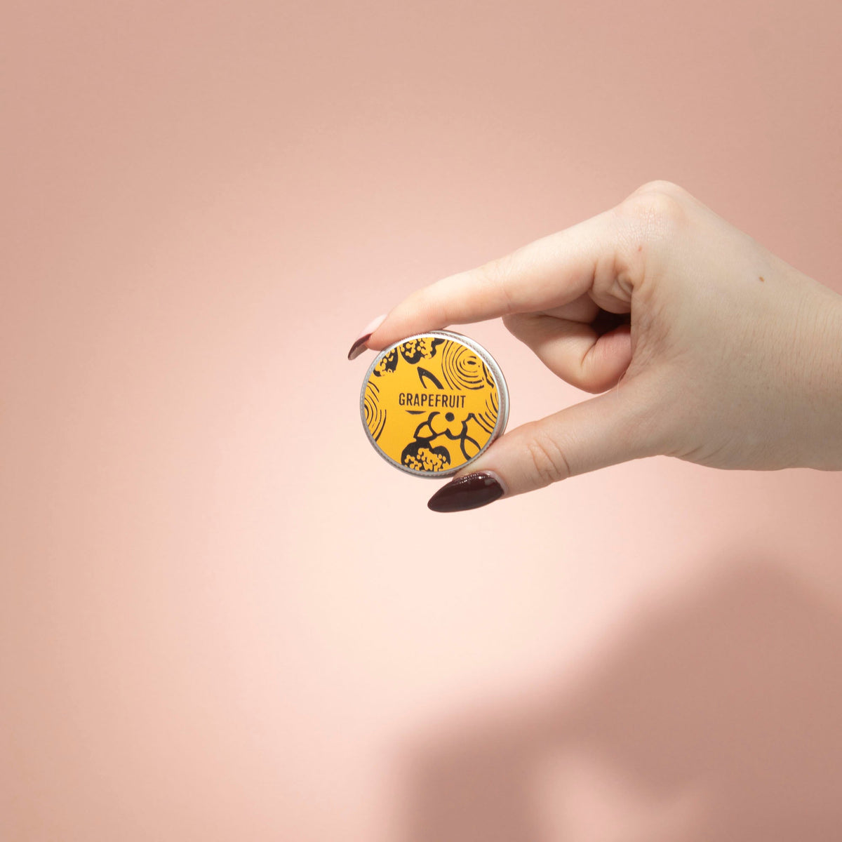 Hand-Poured Vegan-Friendly Natural Lip Balm - Grapefruit, yellow-orange round tin held between two fingers against a peach backdrop, by Quintessential Holistic Health Ltd