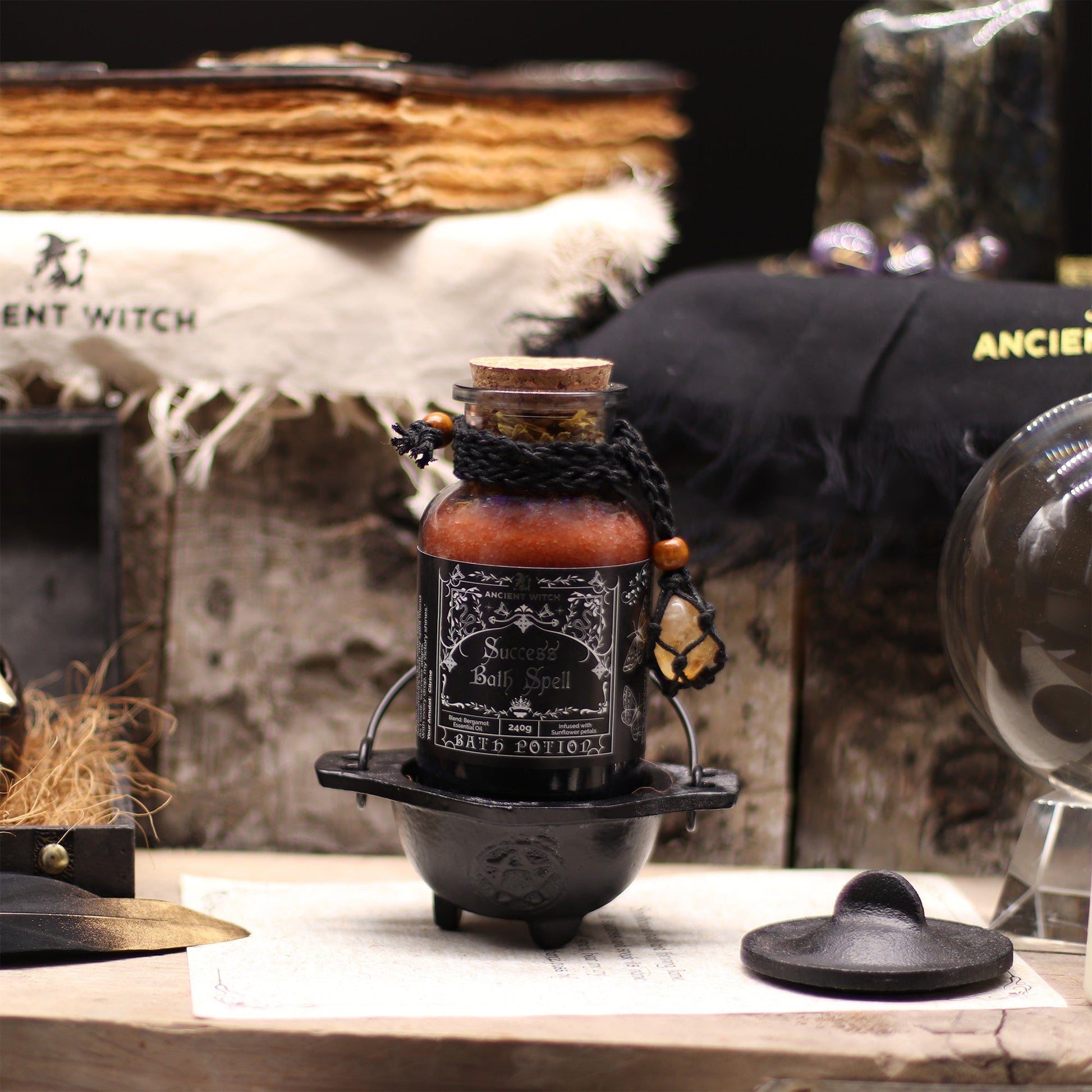 Vegan Bath Spell Potion & Citrine Crystal Amulet - Success, displayed upright in a small black cauldron among books and dark objects, by Quintessential Holistic Health Ltd