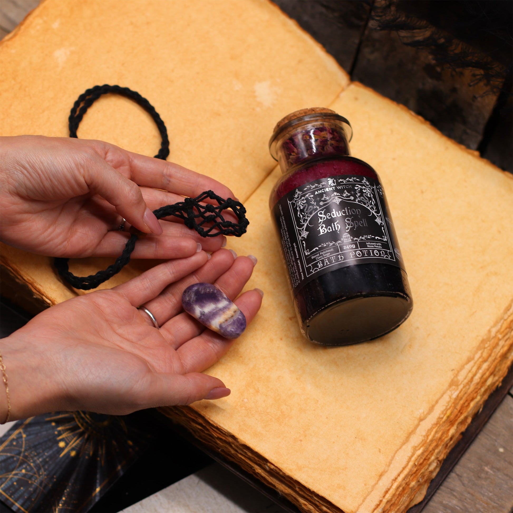 Vegan Bath Spell Potion & Citrine Crystal Amulet - Success, bottle beside a black cord and purple stones held over an open book, by Quintessential Holistic Health Ltd
