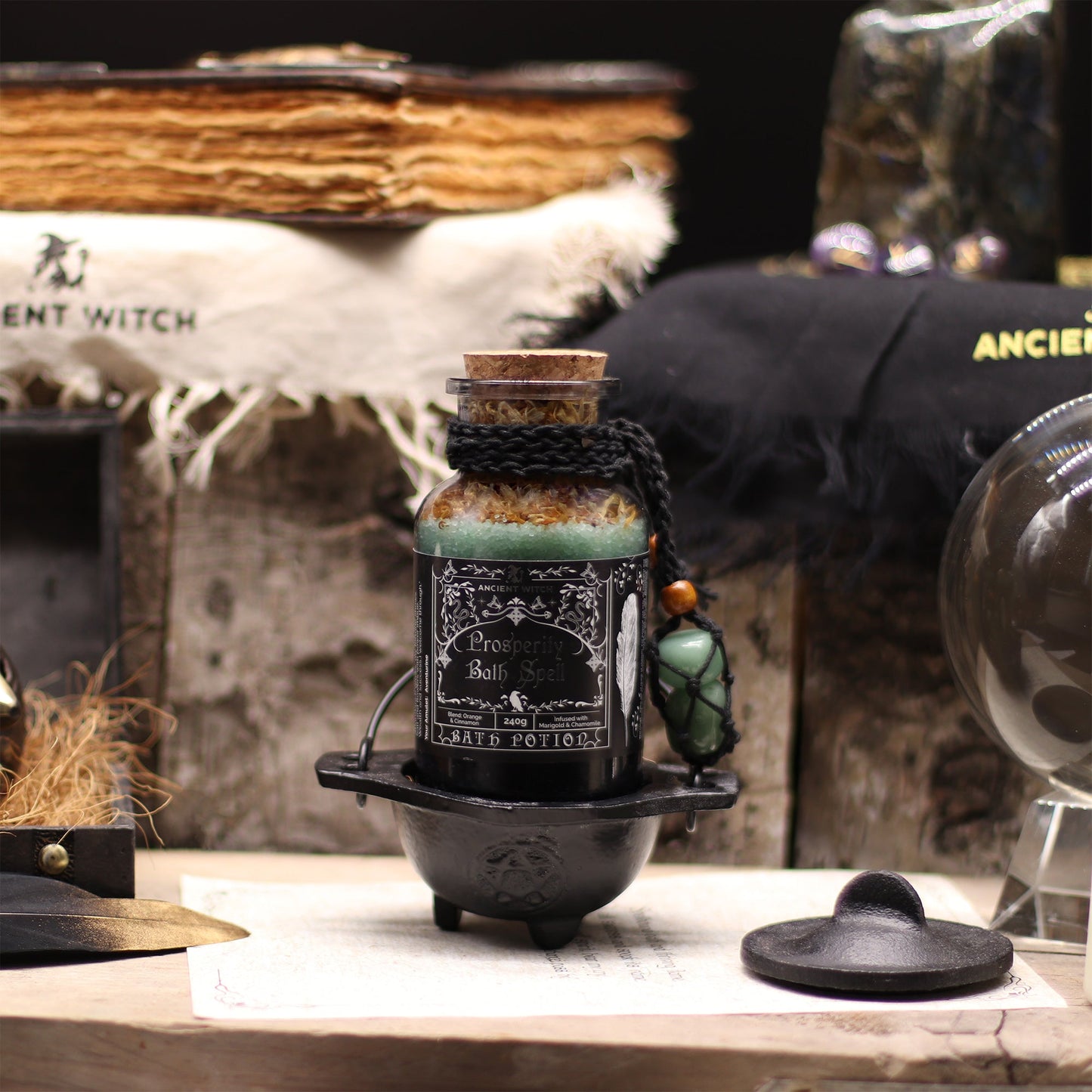 Vegan Bath Spell Potion & Aventurine Crystal Amulet - Prosperity, displayed upright in a small black cauldron among books and dark objects, by Quintessential Holistic Health Ltd