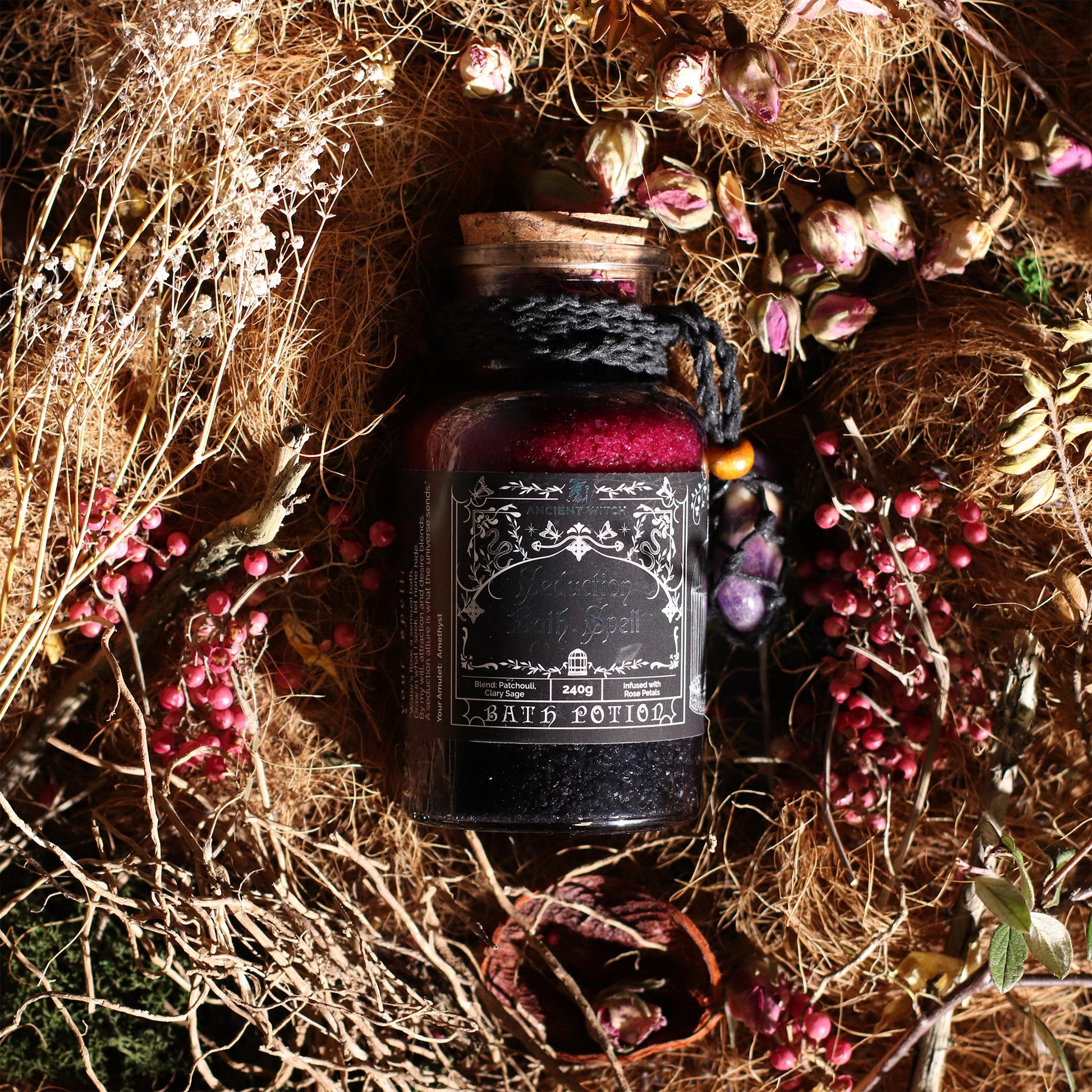 Vegan Bath Spell Potion & Amethyst Crystal Amulet - Seduction, bottle nestled among dry grasses, flowers and scattered pink petals, by Quintessential Holistic Health Ltd