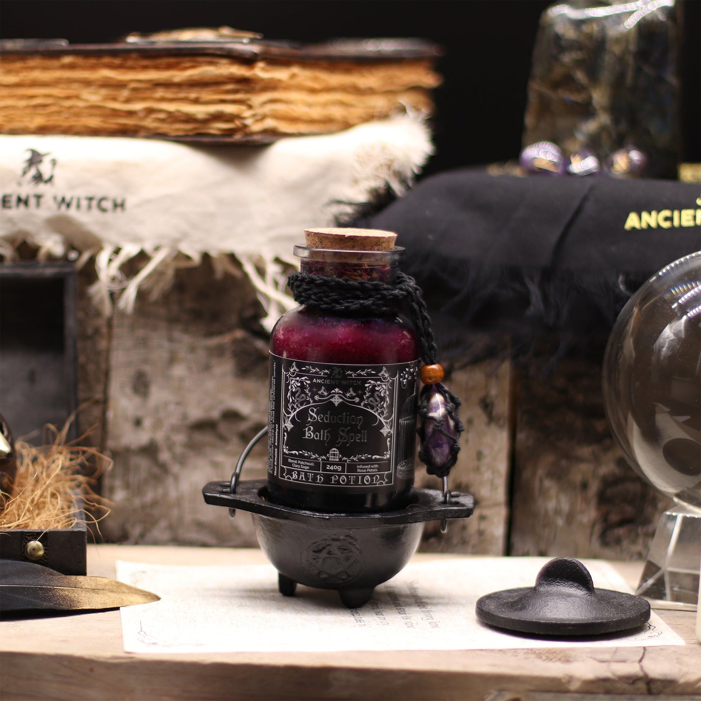 Vegan Bath Spell Potion & Amethyst Crystal Amulet - Seduction, bottle displayed in a small black cauldron among books and dark objects, by Quintessential Holistic Health Ltd