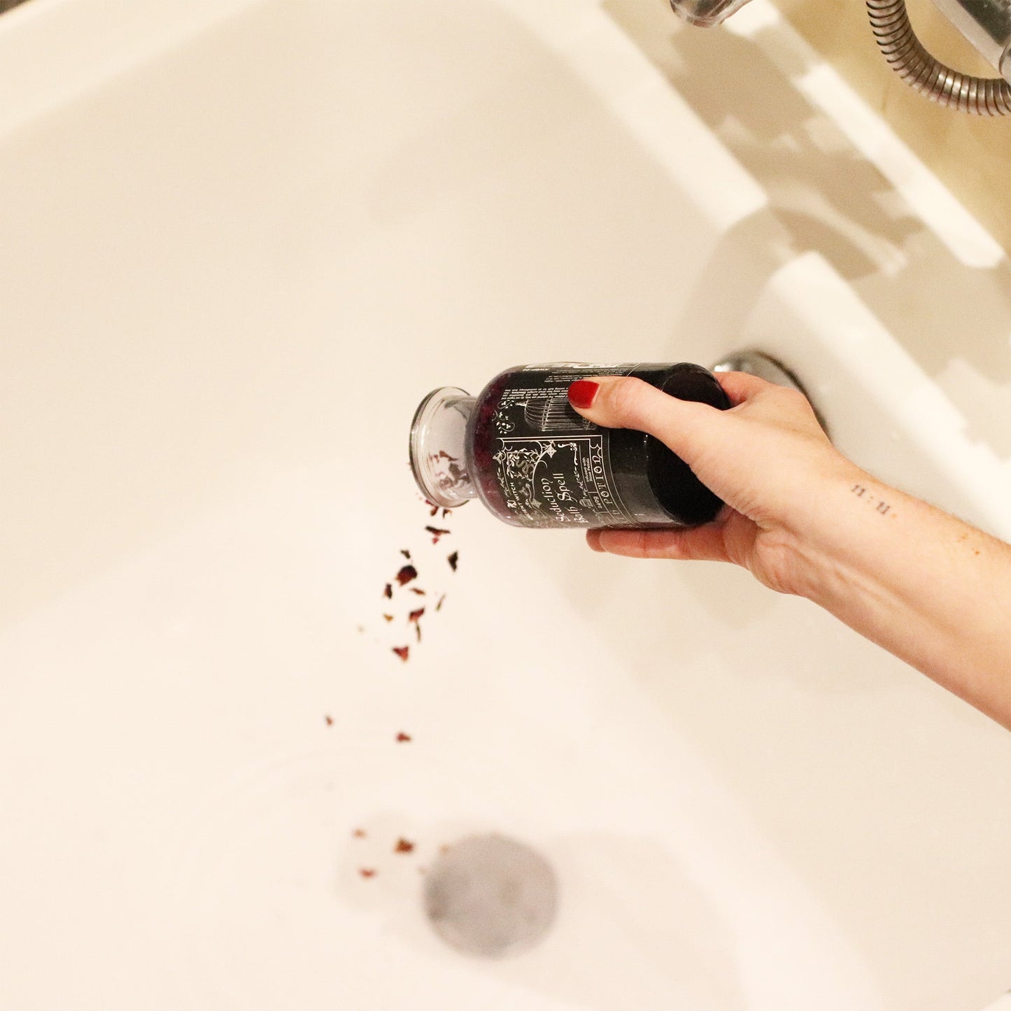 Vegan Bath Spell Potion & Amethyst Crystal Amulet - Seduction, bottle tipped by a hand with red nails as pink petals fall into a white bath, by Quintessential Holistic Health Ltd
