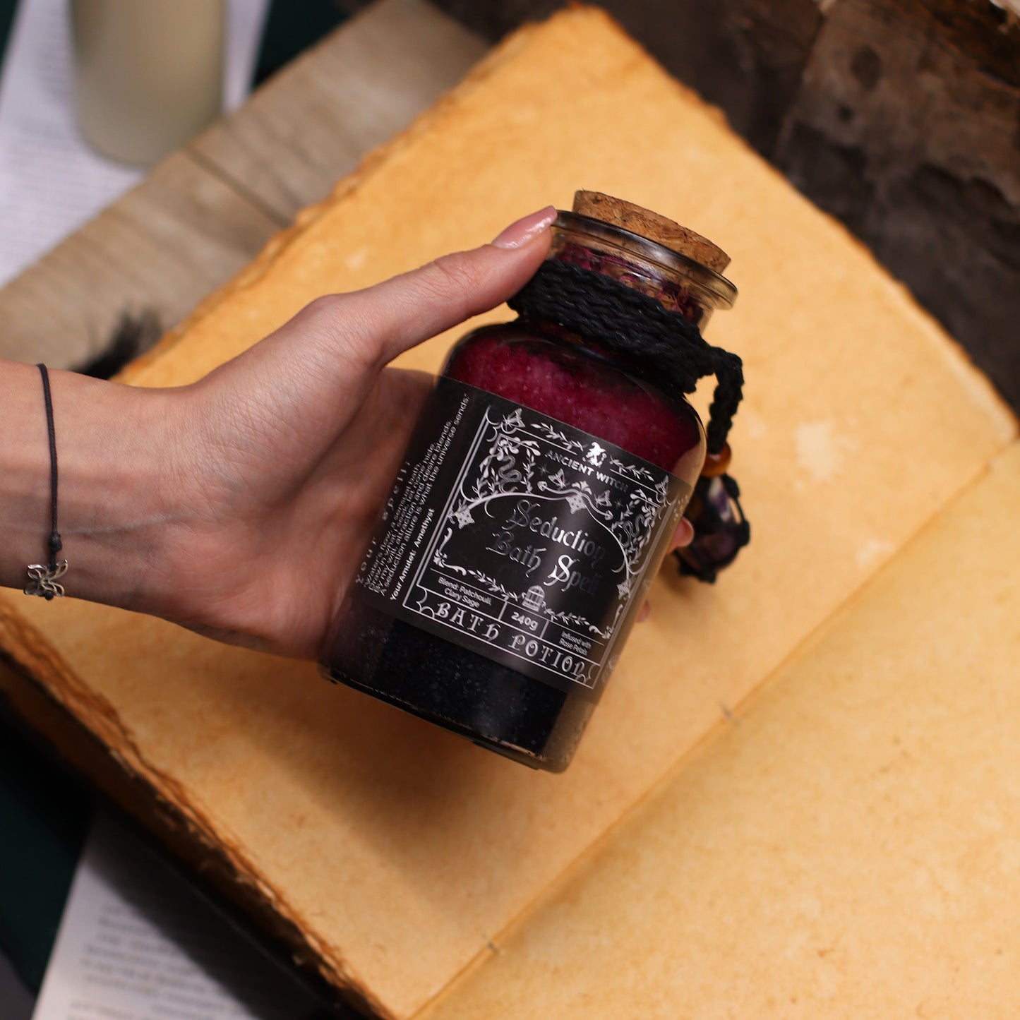Vegan Bath Spell Potion & Amethyst Crystal Amulet - Seduction, labelled bottle held in one hand above an open book, by Quintessential Holistic Health Ltd