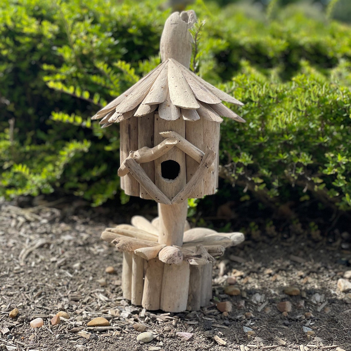 Handmade Recycled Driftwood Bird Box on a Stand, displayed outdoors with a layered roof, round entrance and log base, by Quintessential Holistic Health Ltd