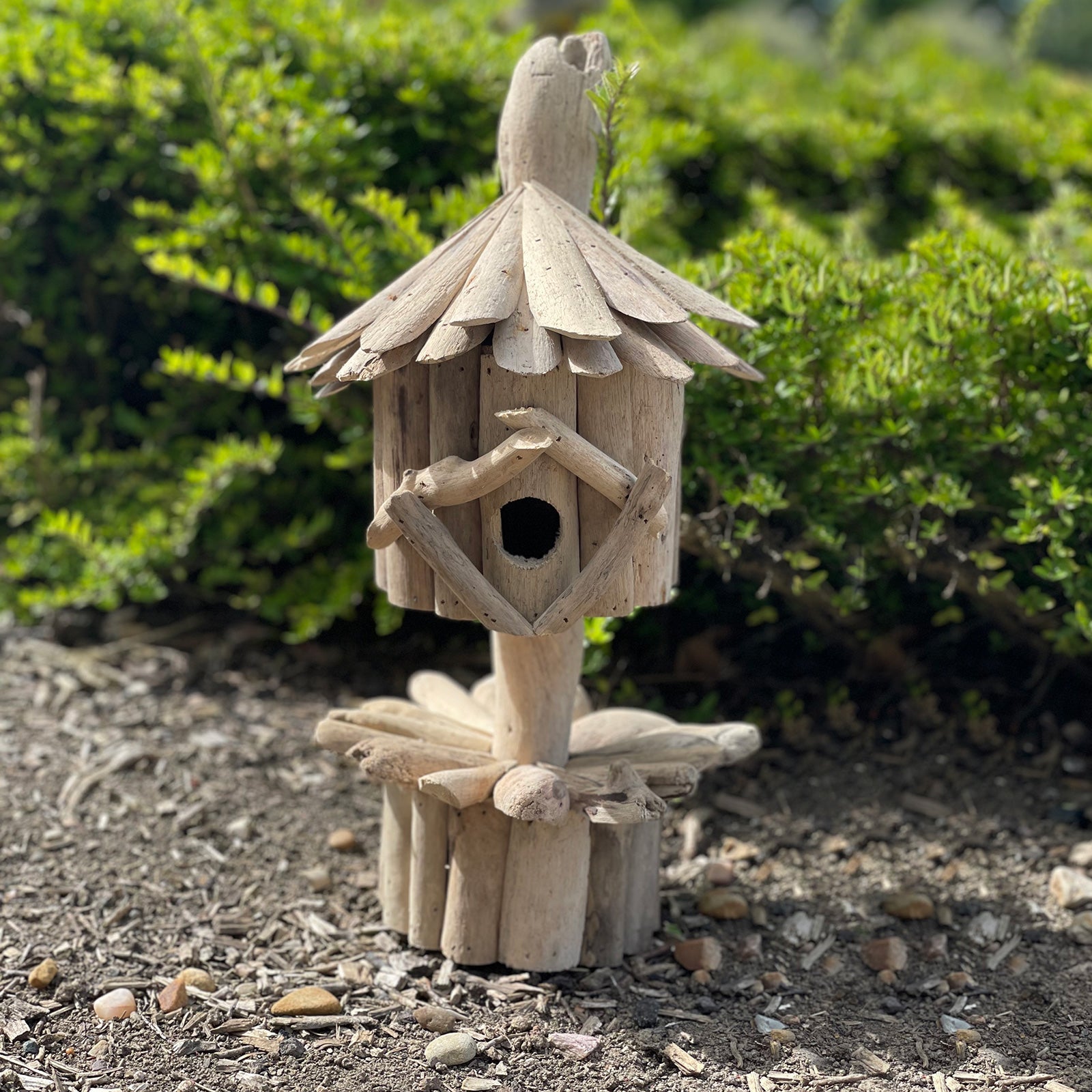 Handmade Recycled Driftwood Bird Box on a Stand, displayed outdoors with a layered roof, round entrance and log base, by Quintessential Holistic Health Ltd