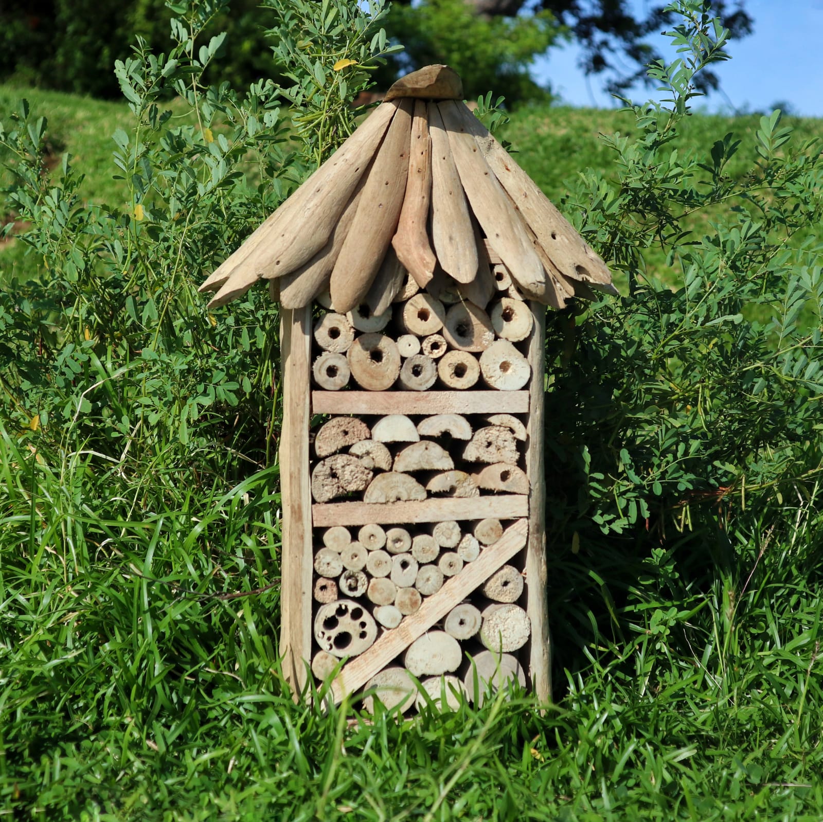 Handmade Recycled Driftwood Bee and Insect Tall House, displayed outdoors with filled sections, a diagonal brace and pitched roof, by Quintessential Holistic Health Ltd