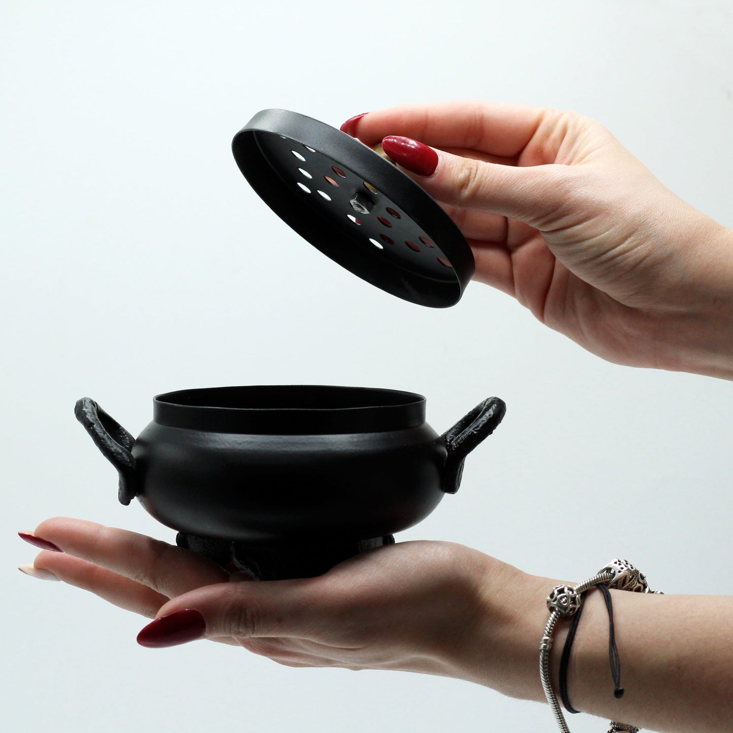 Cast Iron Incense Burner - Classic Cauldron, small black pot resting on a palm while its perforated lid is lifted by Quintessential Holistic Health Ltd
