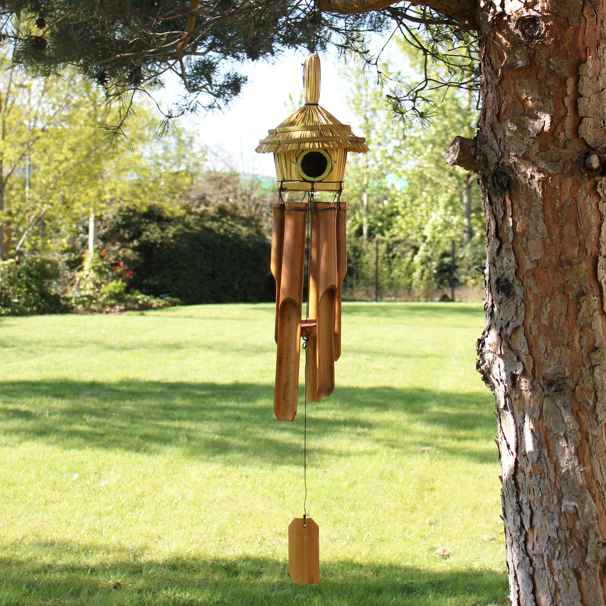 Handcrafted Seagrass & Bamboo Wind Chime Bird Box - Medium Round, hanging from a tree with brown tubes and a rectangular clapper, by Quintessential Holistic Health Ltd