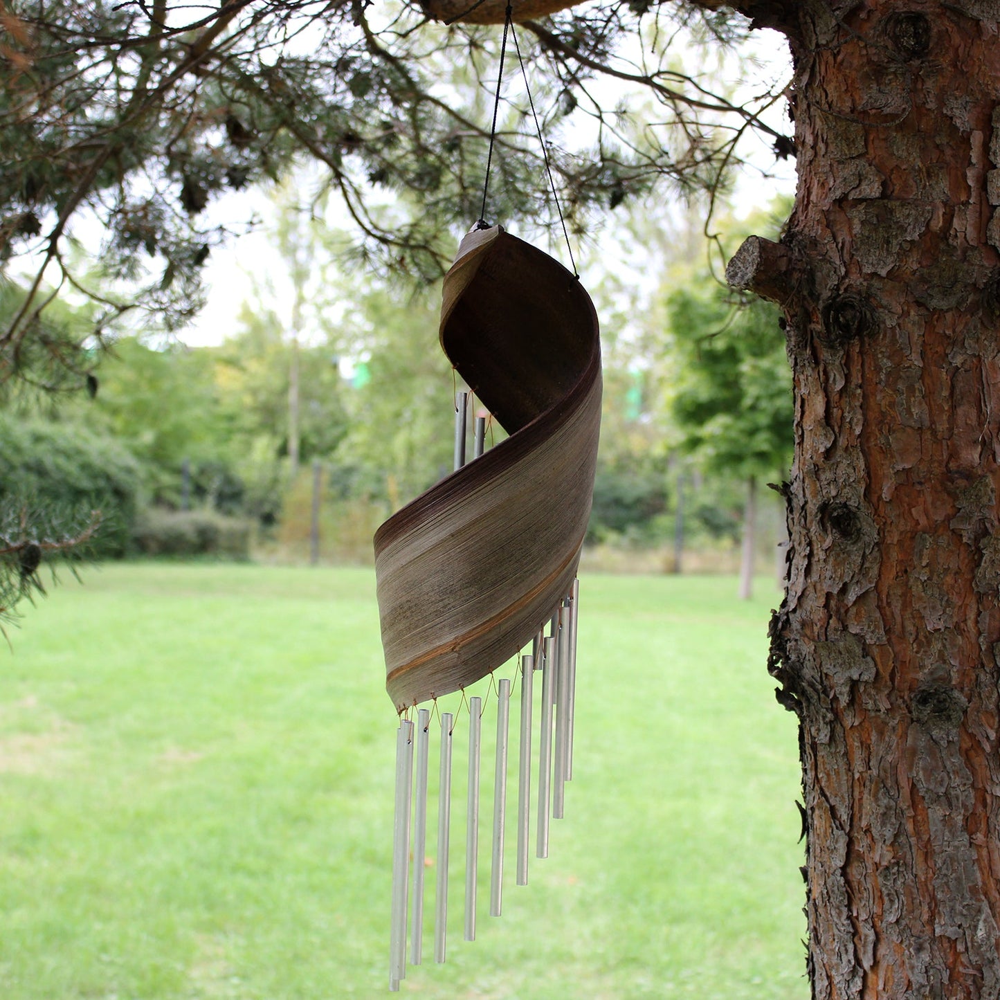 Handcrafted Coconut Leaf Wind Chime - Natural beside a tree with a sweeping brown leaf and hanging silver-tone tubes by Quintessential Holistic Health Ltd