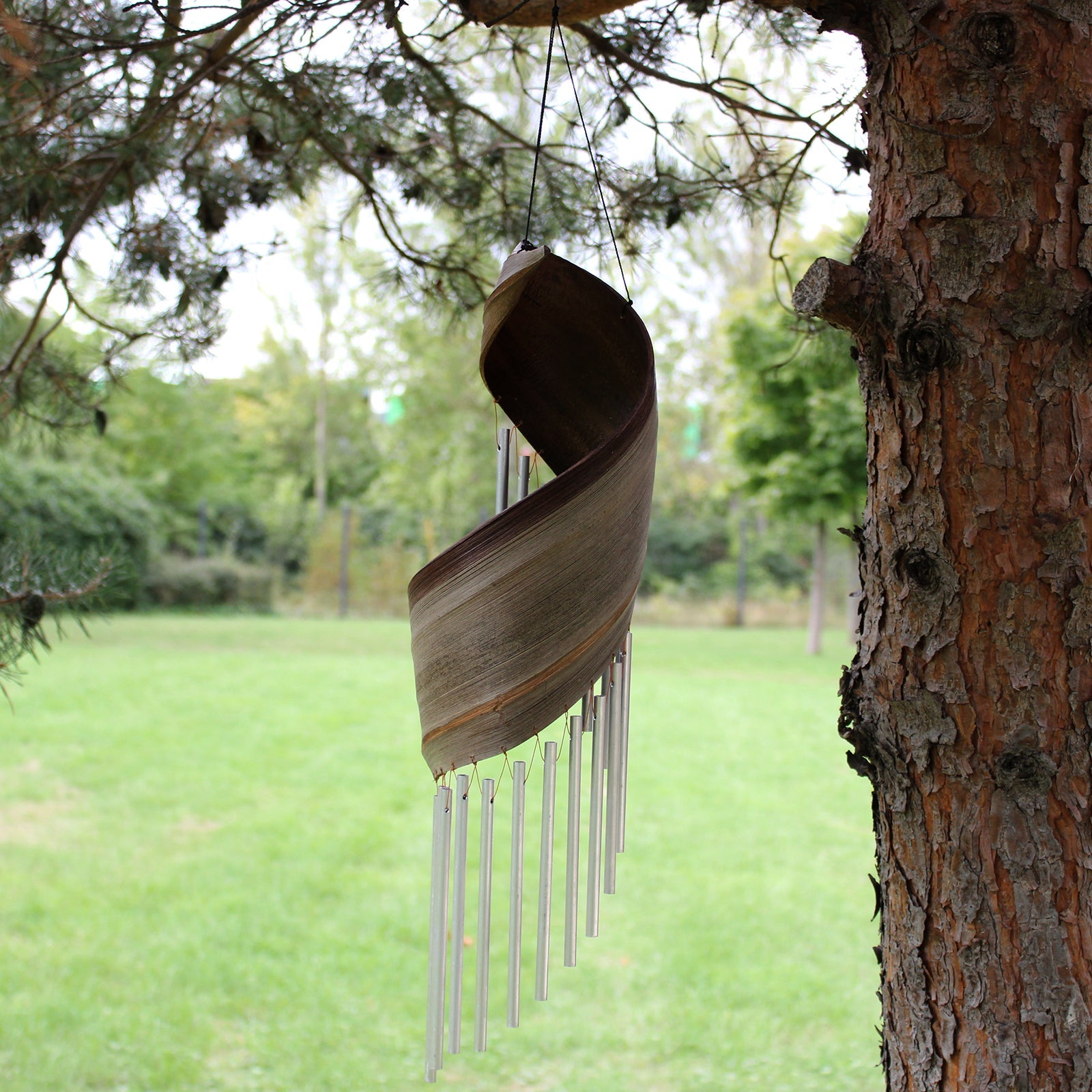 Handcrafted Coconut Leaf Wind Chime - Natural beside a tree with a sweeping brown leaf and hanging silver-tone tubes by Quintessential Holistic Health Ltd