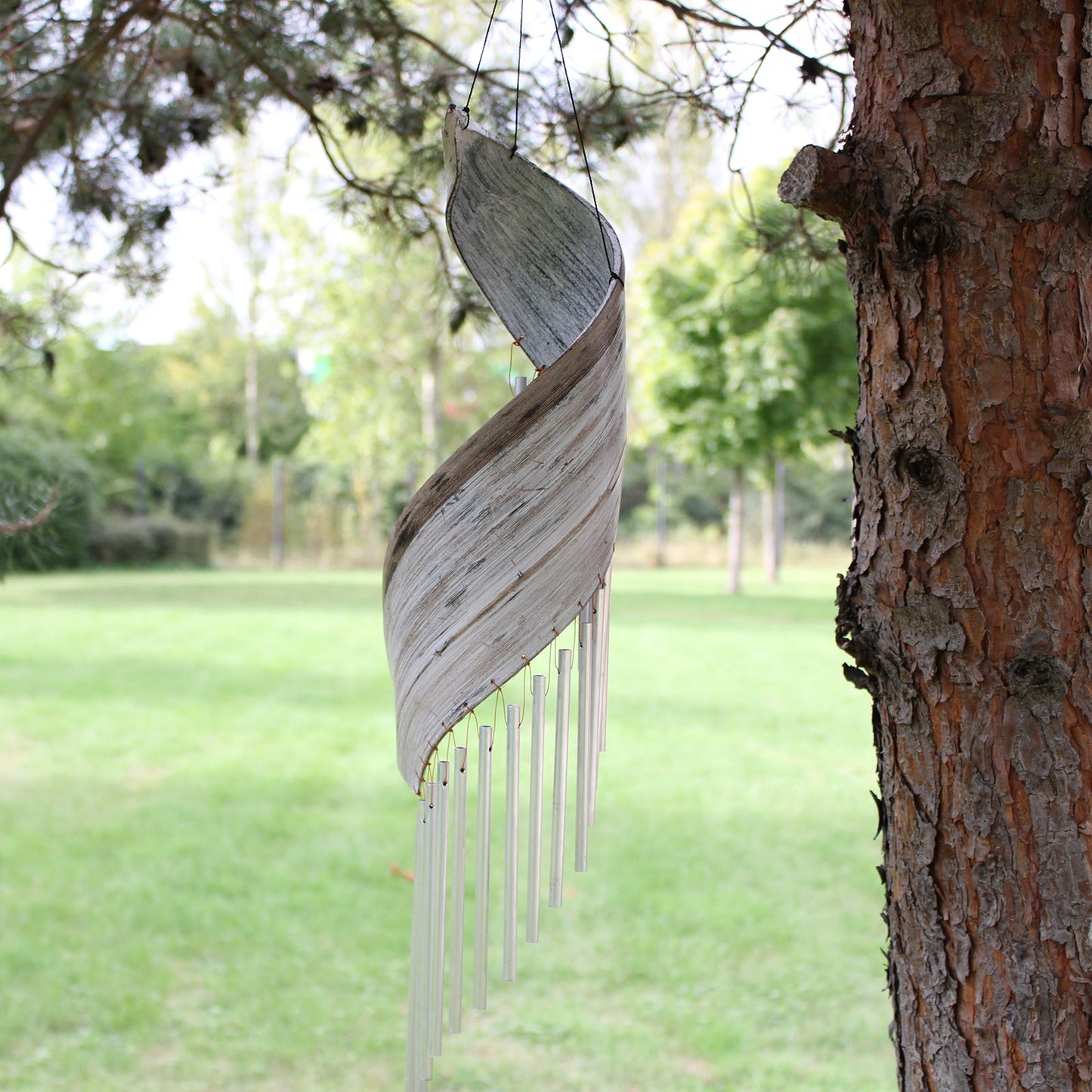 Handcrafted Coconut Leaf Wind Chime - Whitewash hanging beside a tree with a pale grey spiral leaf and silver-tone tubes by Quintessential Holistic Health Ltd