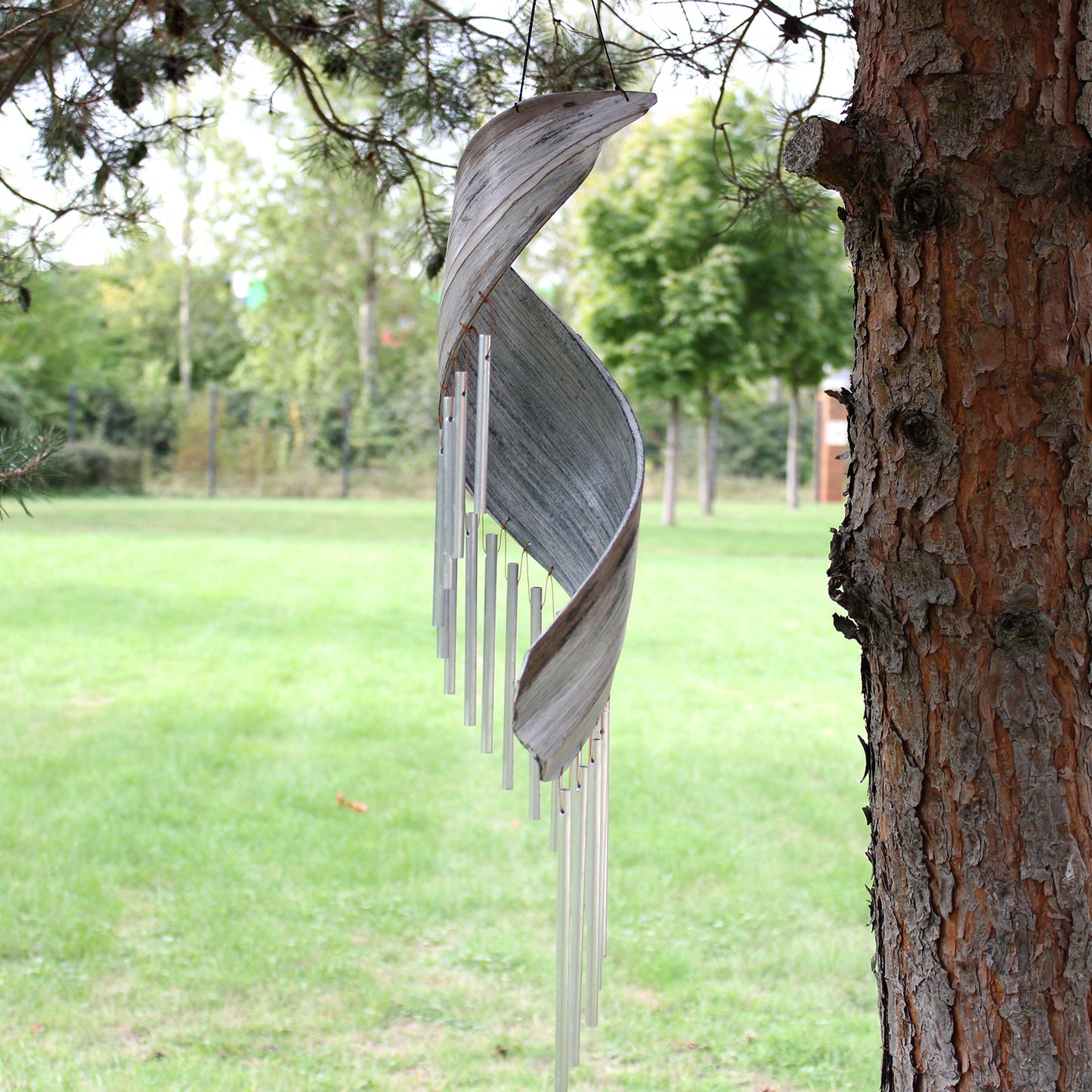 Handcrafted Coconut Leaf Wind Chime - Whitewash beside a tree with a slim pale spiralling profile and descending silver-tone tubes by Quintessential Holistic Health Ltd
