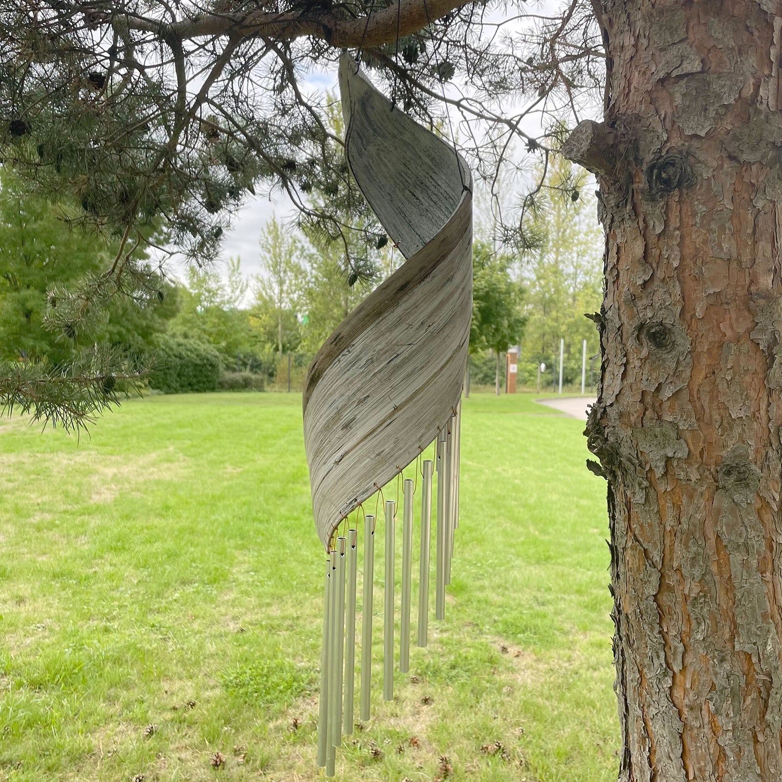Handcrafted Coconut Leaf Wind Chime - Whitewash hanging near a tree with overlapping pale leaf panels and silver-tone tubes by Quintessential Holistic Health Ltd