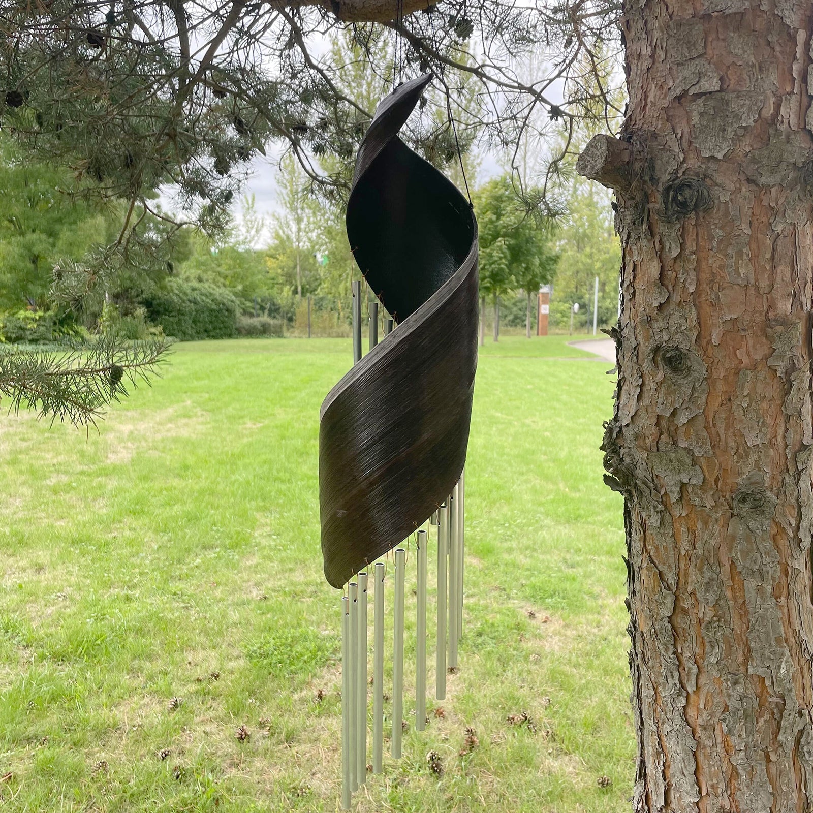 Handcrafted Coconut Leaf Wind Chime - Chocolate hanging beside a tree with a spiralled dark leaf and silver-tone tubes by Quintessential Holistic Health Ltd