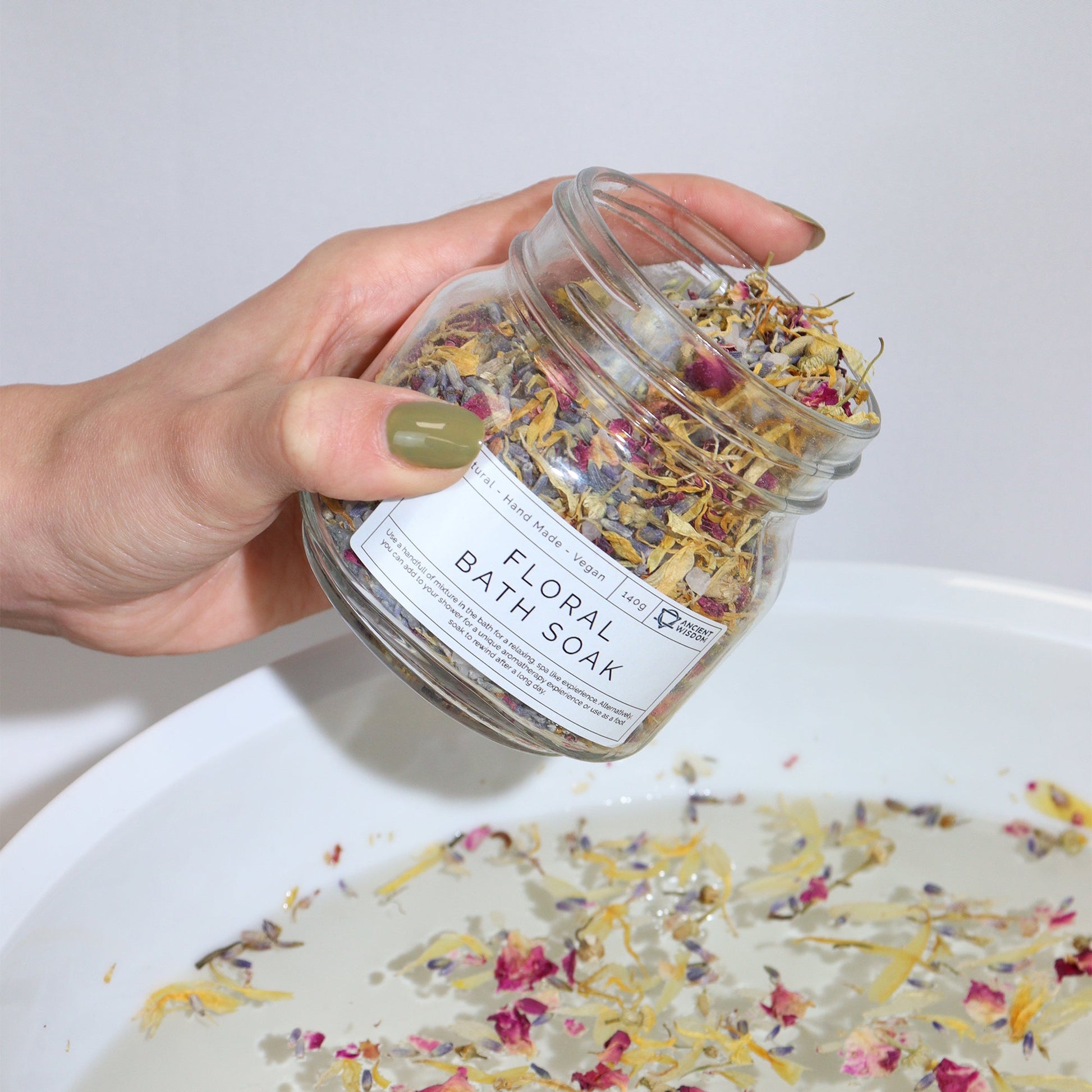 Natural Floral Bath Soak - 140g, hand holding a labelled jar above a white bowl scattered with colourful petals, by Quintessential Holistic Health Ltd