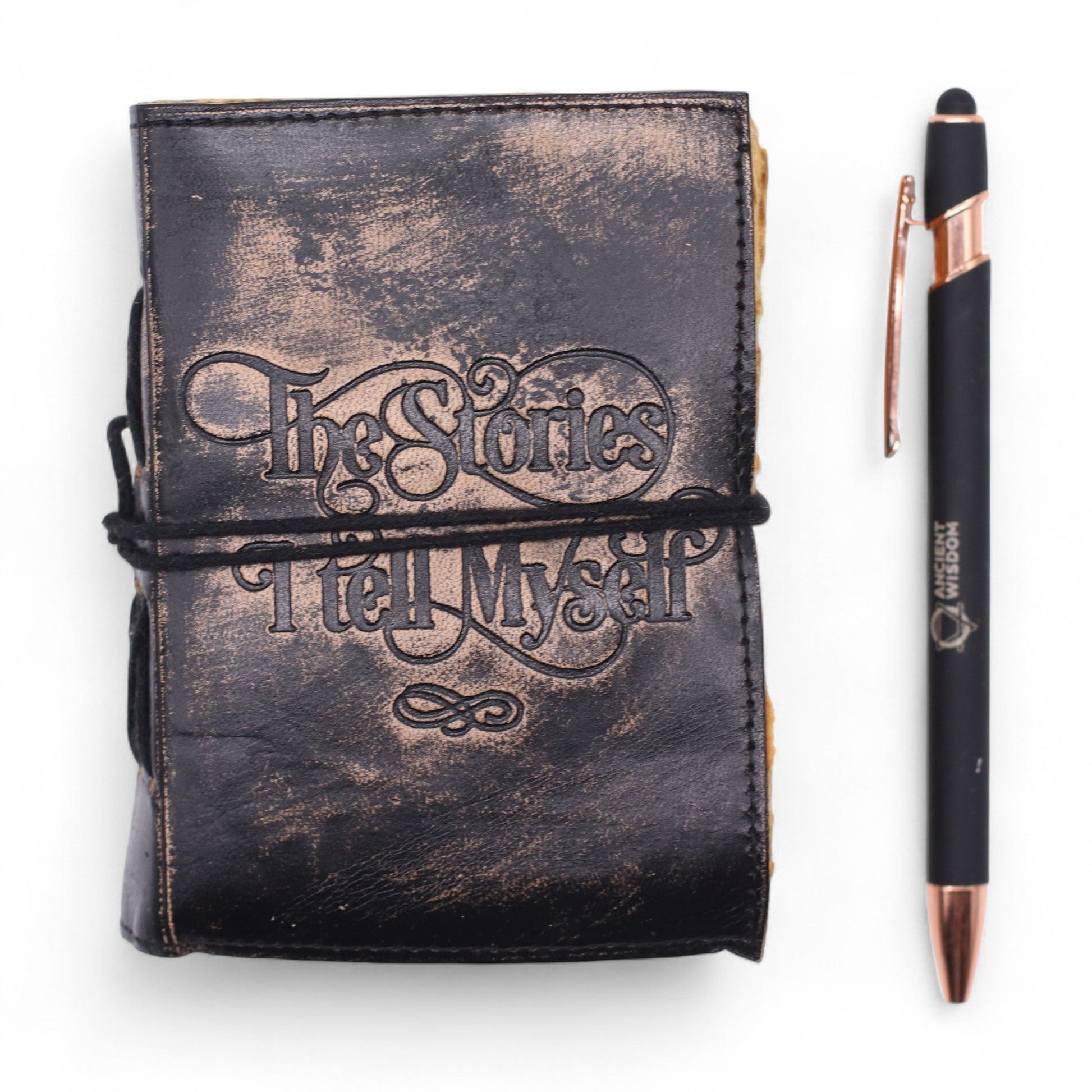 Small Black and Tan Leather Journal - The Stories I Tell Myself with ornate cover lettering and wrap cord beside a black pen by Quintessential Holistic Health Ltd