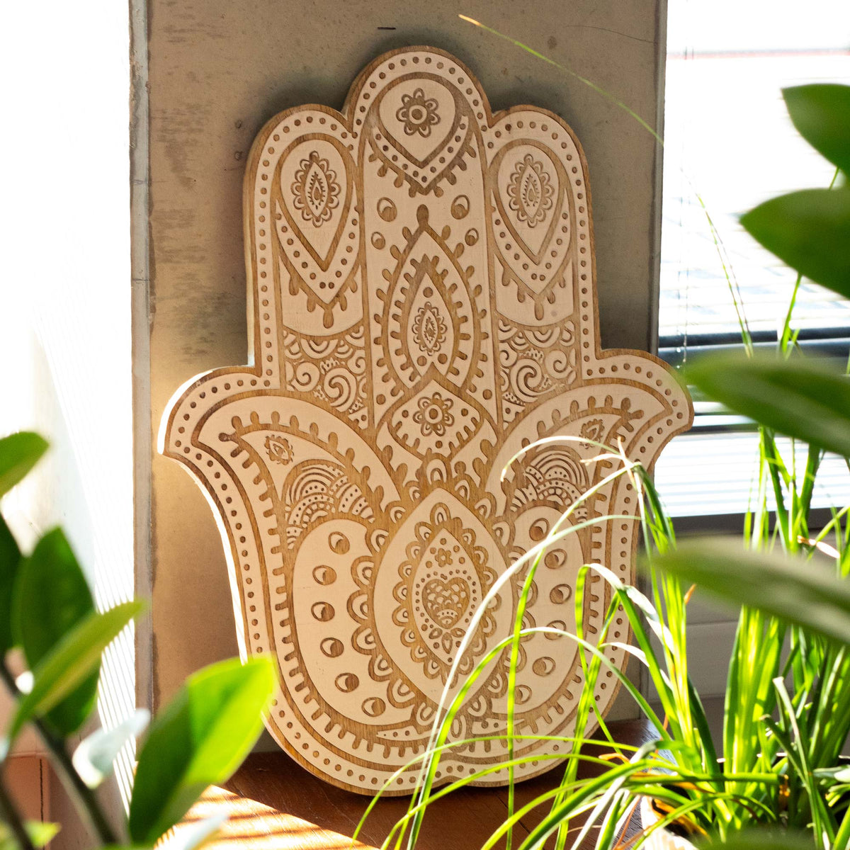 Hand-Carved Mango Wood Hamsa Wall Plaque - Whitewash, standing upright among green leaves with its carved face visible, by Quintessential Holistic Health Ltd