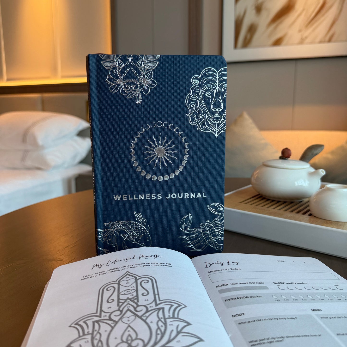 Wellness Journal - A5 - Midnight Blue and Silver Zodiac, closed journal standing behind open illustrated pages in a softly lit room by Quintessential Holistic Health Ltd
