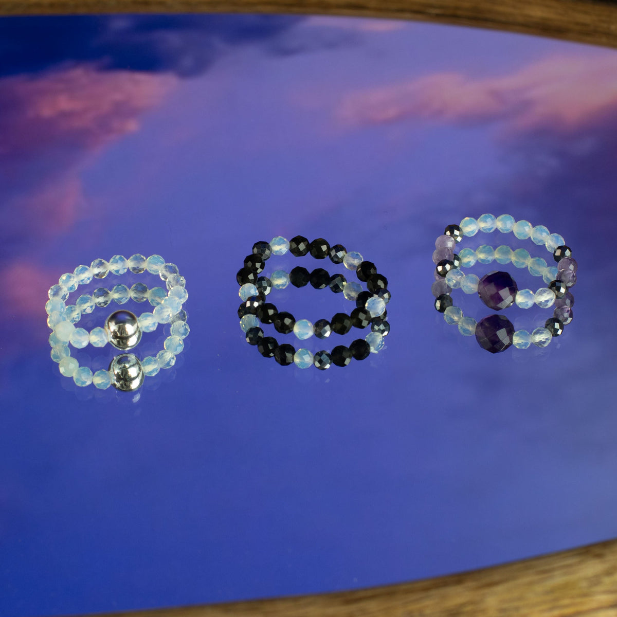 Mystical Nymph Spirit Gemstone 3-Ring Set – Full Moon, three pale, black and purple beaded rings arranged on a glossy blue and purple surface, by Quintessential Holistic Health Ltd