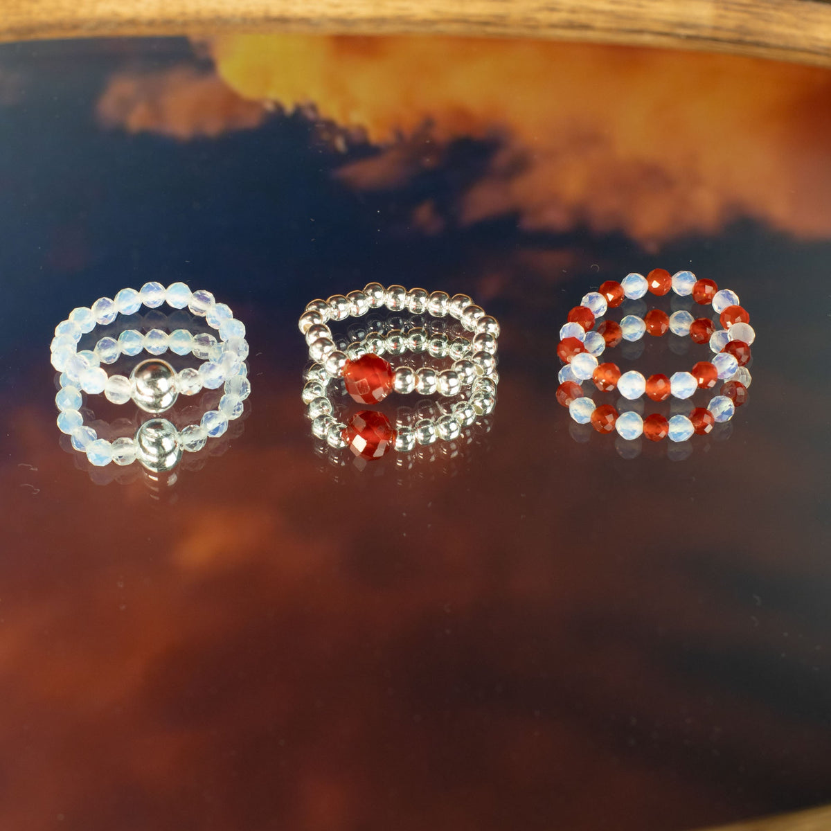 Mystical Nymph Spirit Gemstone 3-Ring Set – Blood Moon, three beaded rings arranged separately on a reflective amber surface by Quintessential Holistic Health Ltd