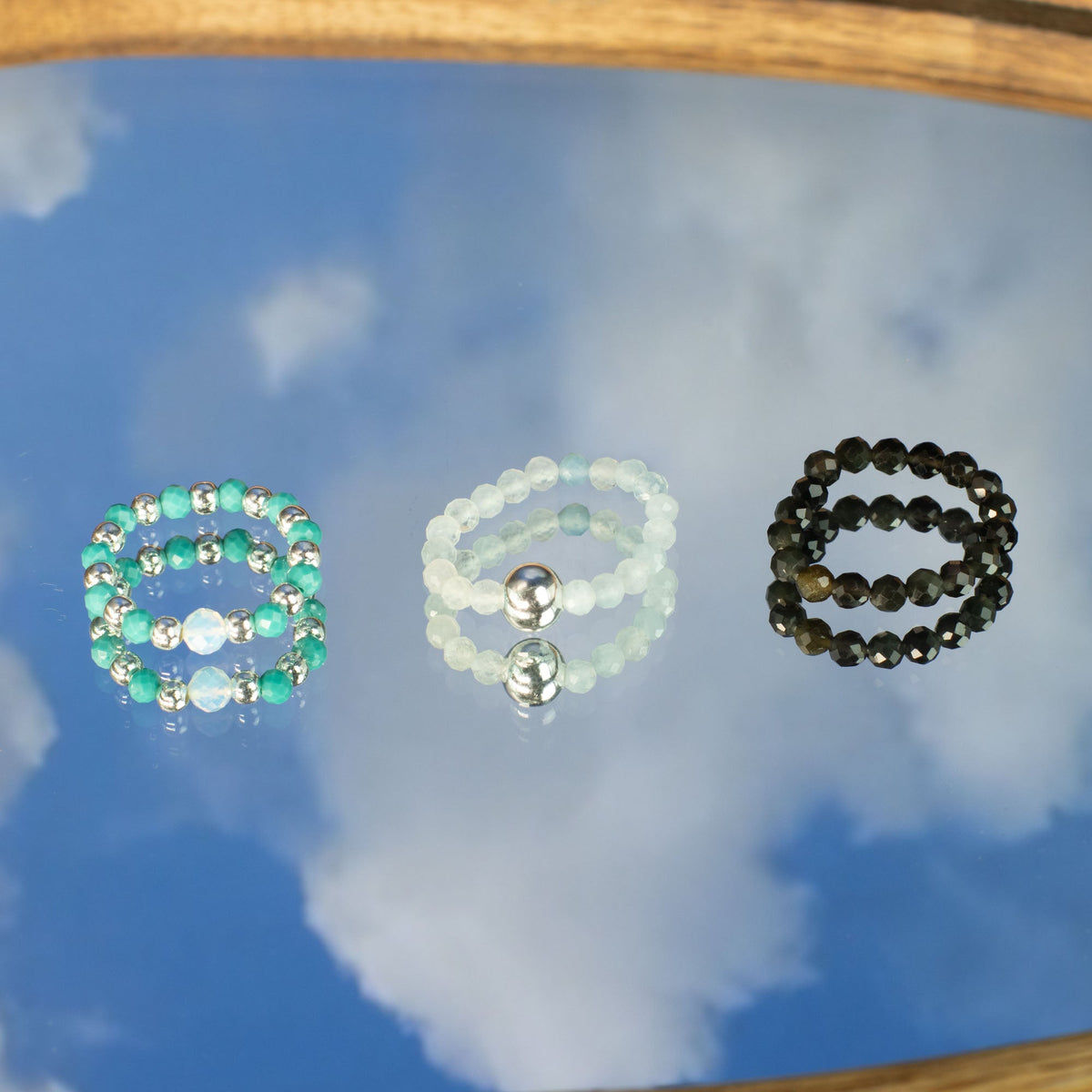 Mystical Nymph Spirit Gemstone 3-Ring Set – Endless Ocean, three beaded rings arranged on a reflective sky-and-cloud surface by Quintessential Holistic Health Ltd