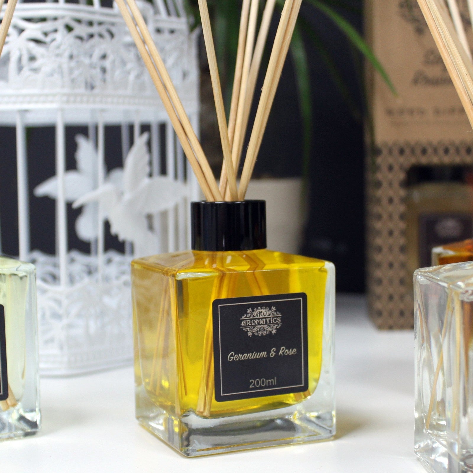 Reed Diffuser - Geranium and Rose - 200ml, close view of an amber-filled square bottle with black label and reeds among other diffusers by Quintessential Holistic Health Ltd