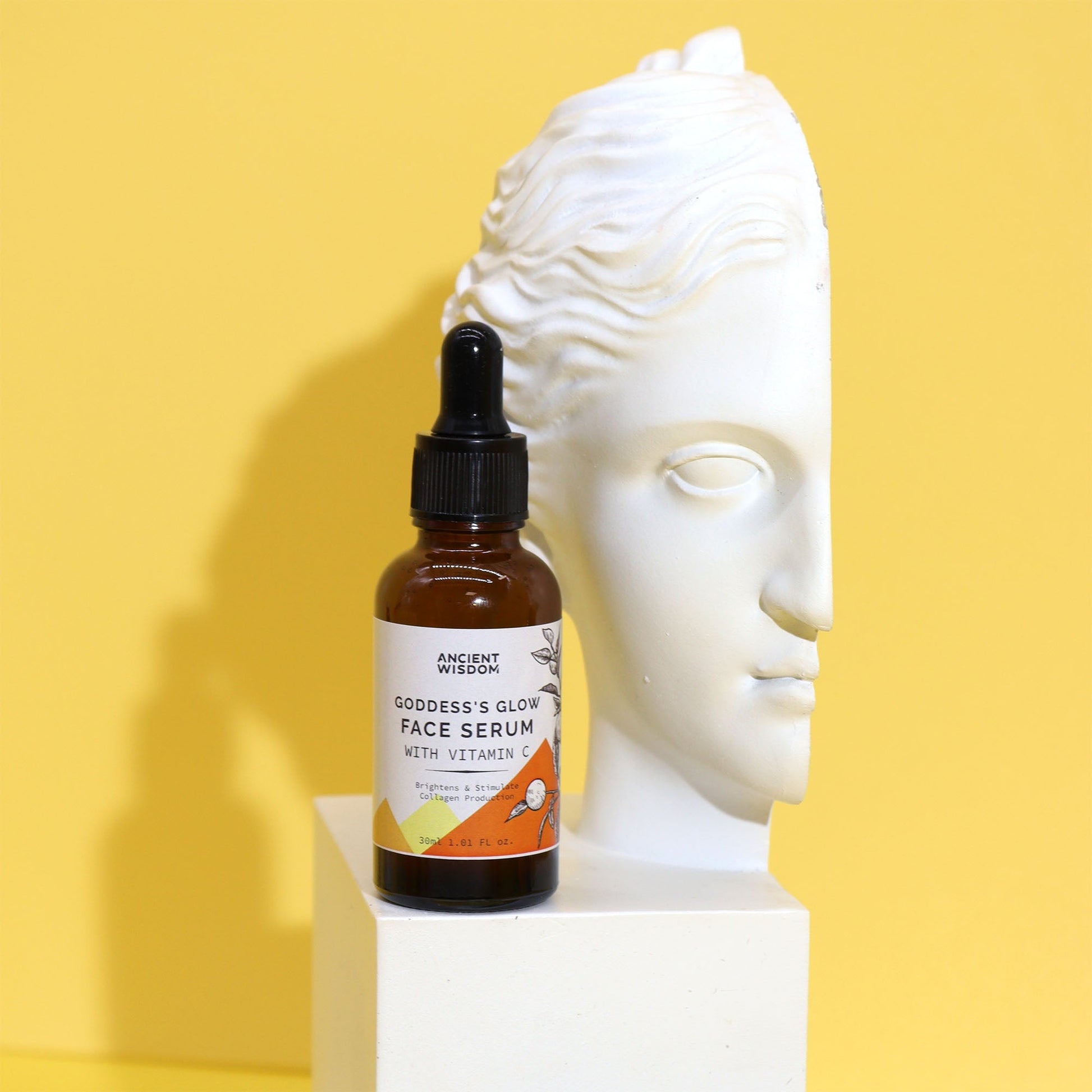 Vitamin C Goddess’s Glow Face Serum in an amber dropper bottle beside a white sculpted head against a yellow background by Quintessential Holistic Health Ltd