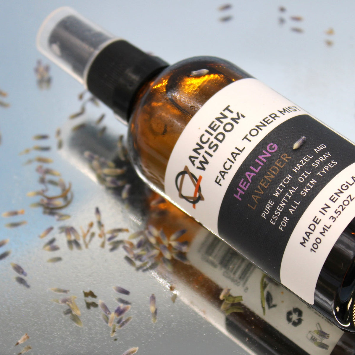 100ml Vegan-Friendly Witch Hazel Facial Toner Mist - Lavender in an amber spray bottle lying beside scattered purple flower sprigs by Quintessential Holistic Health Ltd