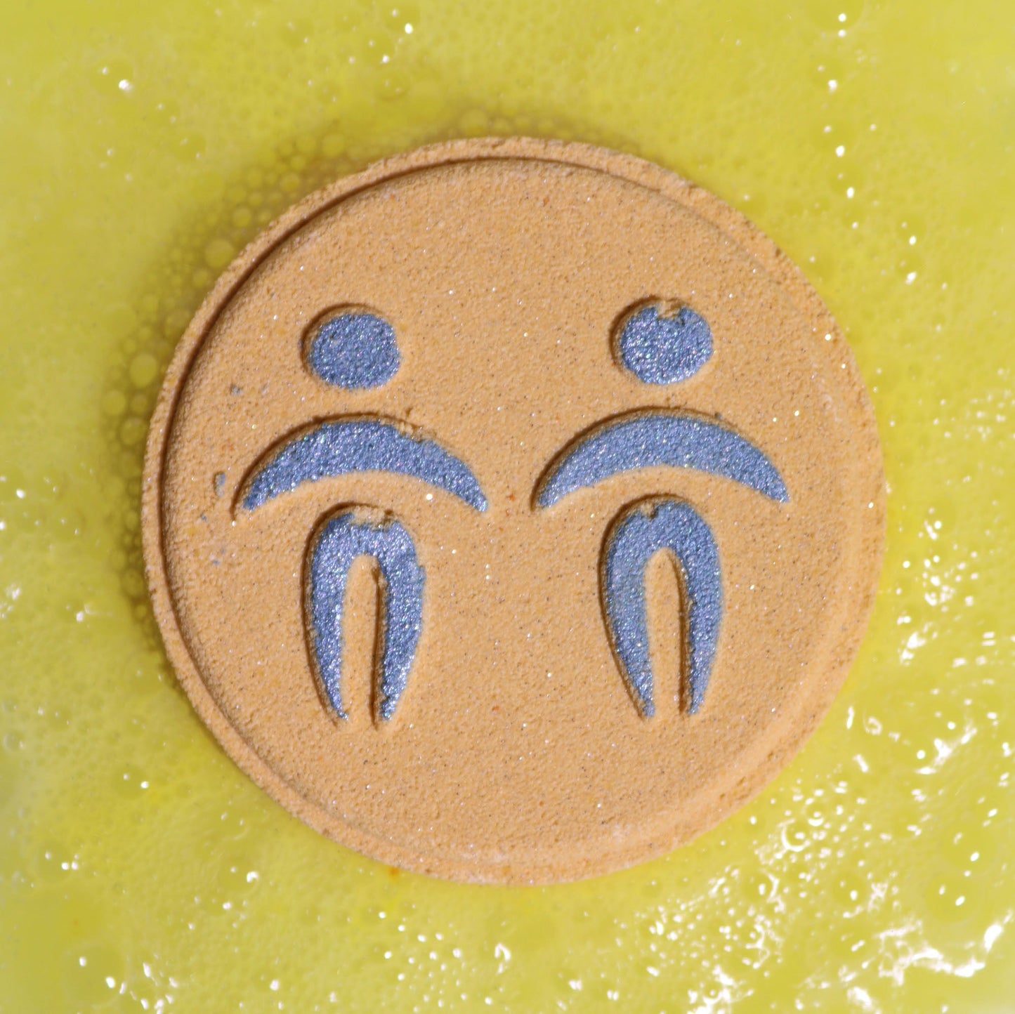 Gemini Bath Bomb Fizzers - Oriental Tigers Eye - 2 Pack, close-up of a yellow round fizzer embossed with the Gemini symbol by Quintessential Holistic Health Ltd